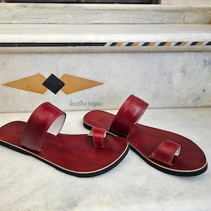 May include: A pair of red leather sandals with a toe loop and a wide strap. The sandals have a black sole and a white interior. The sandals are displayed on a marble surface. The text "leathereyes" is visible.