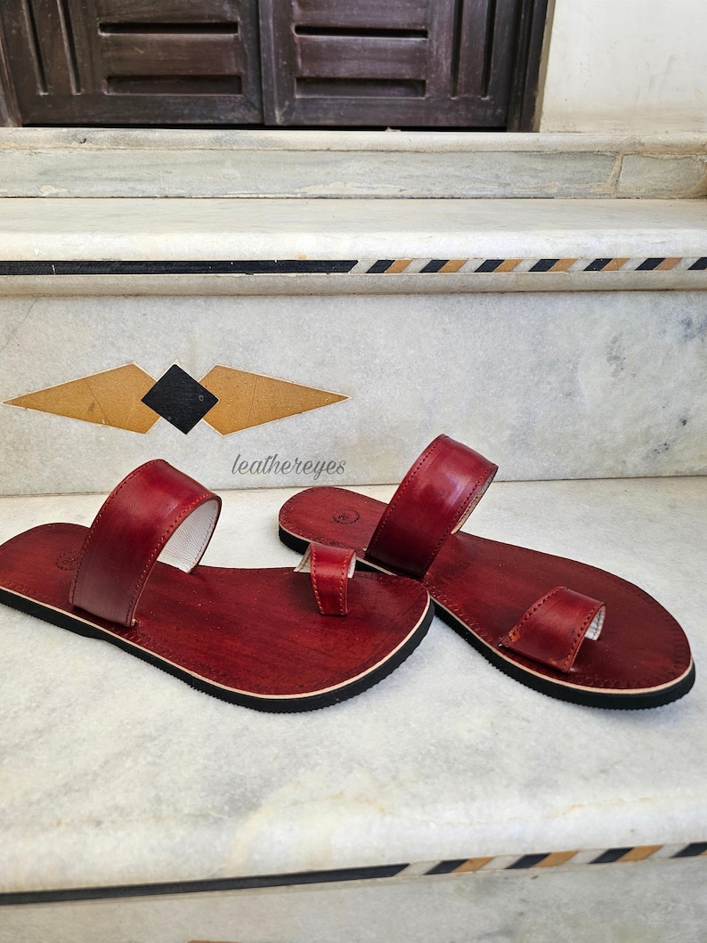 May include: A pair of red leather sandals with a toe loop and a wide strap. The sandals have a black sole and a white interior. The sandals are displayed on a marble surface. The text "leathereyes" is visible.