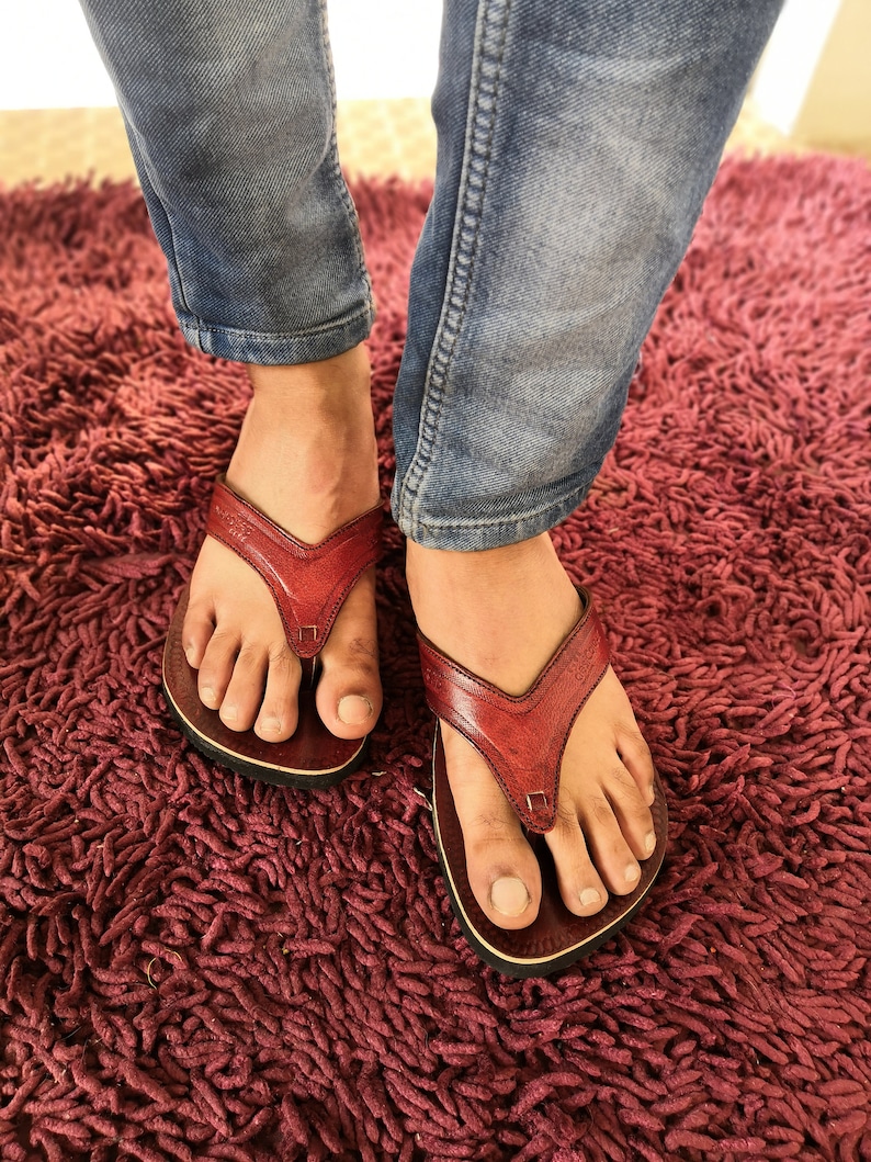May include: A pair of red leather sandals with a simple design. The sandals are worn by a person wearing blue jeans.