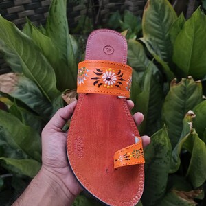 Op de afbeelding: Handgemaakte oranje leren sandaal met bloemmotieven. De sandaal heeft een teengreep en een band versierd met geschilderde bloemen en bladeren. De zool is donkeroranje en de sandaal wordt tegen een achtergrond van groen gebladerte gehouden.