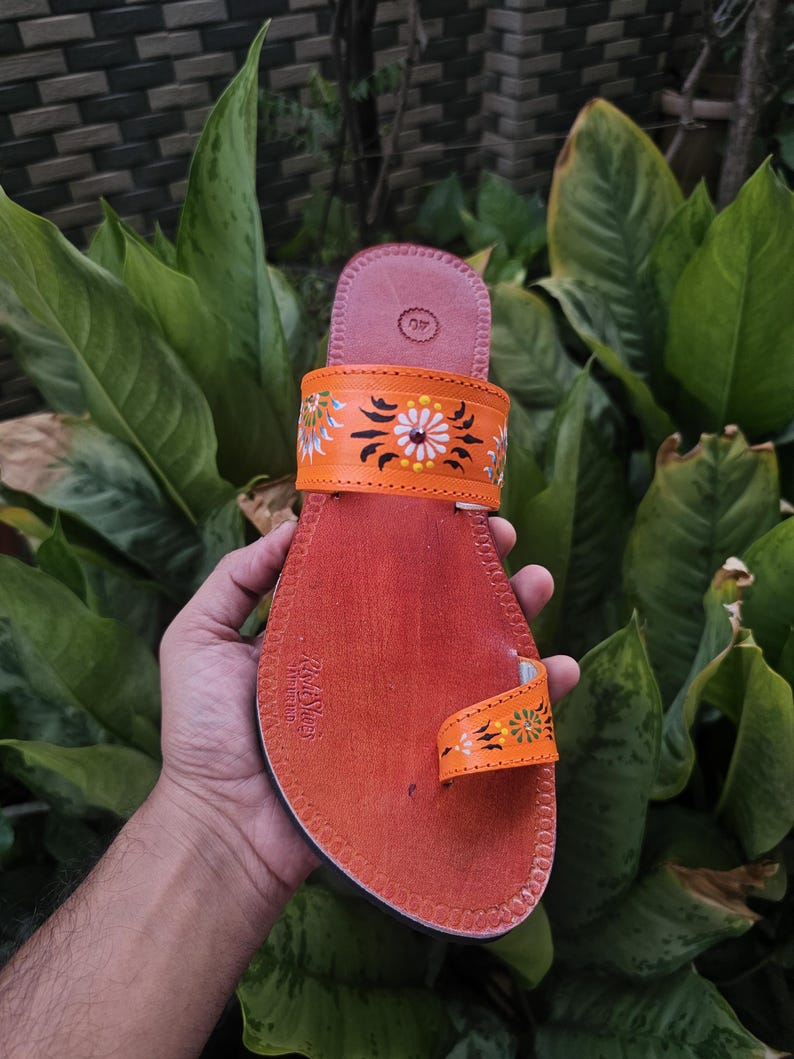 Op de afbeelding: Handgemaakte oranje leren sandaal met bloemmotieven. De sandaal heeft een teengreep en een band versierd met geschilderde bloemen en bladeren. De zool is donkeroranje en de sandaal wordt tegen een achtergrond van groen gebladerte gehouden.
