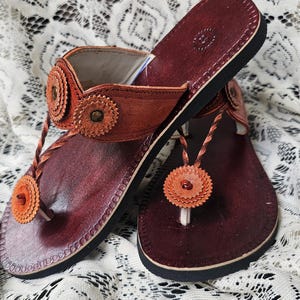 May include: A pair of leather sandals with a dark burgundy footbed and a black sole. The sandals feature a thong strap and decorative orange floral accents. The sandals are displayed on a white lace surface.
