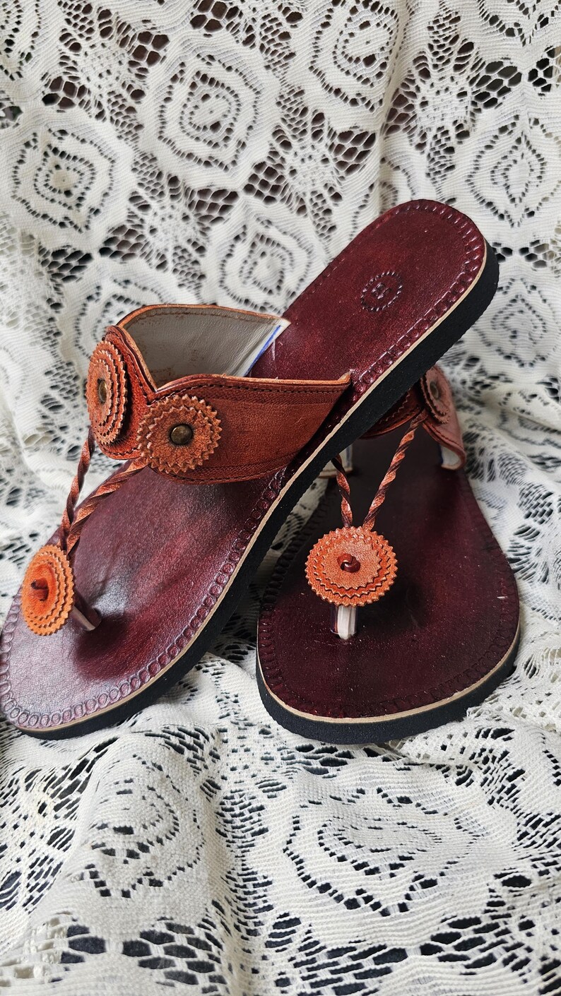 May include: A pair of leather sandals with a dark burgundy footbed and a black sole. The sandals feature a thong strap and decorative orange floral accents. The sandals are displayed on a white lace surface.