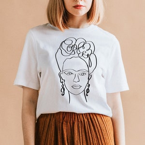 May include: White t-shirt featuring a black line drawing of a woman's face with floral hair and dangling earrings. The design is a minimalist, artistic representation of a famous figure. The shirt is paired with a rust-colored skirt.