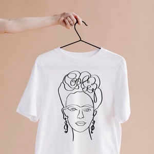 May include: White t-shirt featuring a black line drawing of a woman's face with floral headpiece and earrings. The design is a minimalist, artistic representation. The shirt is hanging on a black hanger against a neutral background.