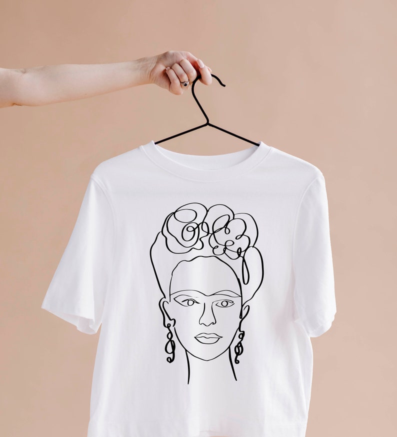 May include: White t-shirt featuring a black line drawing of a woman's face with floral headpiece and earrings. The design is a minimalist, artistic representation. The shirt is hanging on a black hanger against a neutral background.
