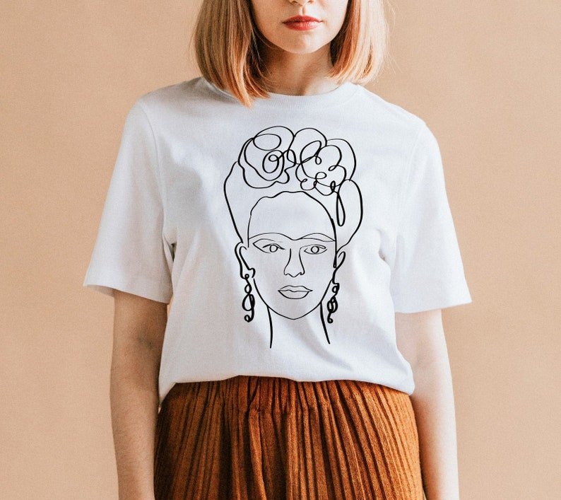 May include: White t-shirt featuring a black line drawing of a woman's face with floral hair and dangling earrings. The design is a minimalist, artistic representation of a famous figure. The shirt is paired with a rust-colored skirt.