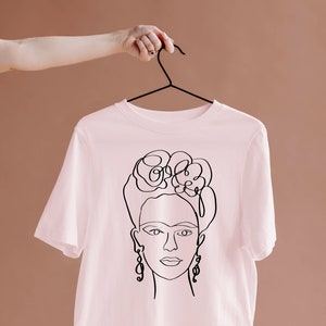 May include: A light pink t-shirt featuring a black line drawing of a woman's face, including her hair, earrings, and facial features. The t-shirt is hanging on a black hanger.