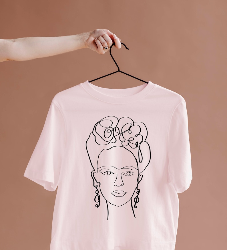 May include: A light pink t-shirt featuring a black line drawing of a woman's face, including her hair, earrings, and facial features. The t-shirt is hanging on a black hanger.