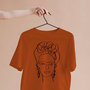 May include: Rust-colored t-shirt featuring a black line drawing of a woman's face with floral hair and dangling earrings. The design is a minimalist, artistic representation. The shirt is hanging on a black hanger.