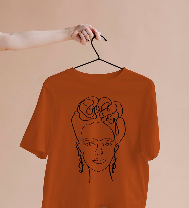 May include: Rust-colored t-shirt featuring a black line drawing of a woman's face with floral hair and dangling earrings. The design is a minimalist, artistic representation. The shirt is hanging on a black hanger.