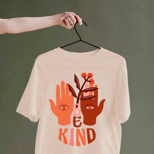 May include: Cream-colored t-shirt with a graphic design featuring two hands, a plant, and the words "BE KIND" in orange and pink. The design has a retro, bohemian aesthetic. The shirt is hanging on a black hanger.