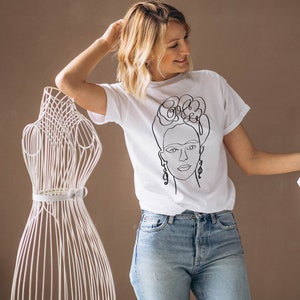May include: White t-shirt featuring a black line drawing of a woman's face with floral headpiece and earrings. The shirt is paired with blue jeans. A white wire dress form is in the background.