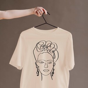May include: A cream-colored t-shirt featuring a black line drawing of a woman's face with floral hair and dangling earrings. The t-shirt is hanging on a black hanger against a neutral background.