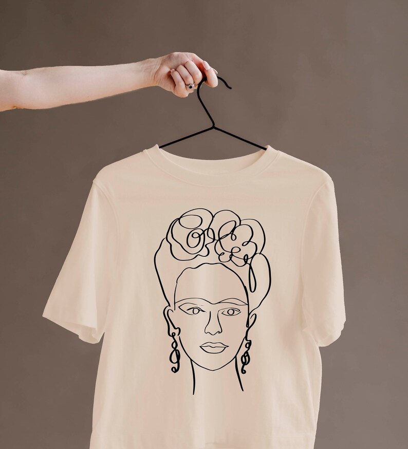 May include: A cream-colored t-shirt featuring a black line drawing of a woman's face with floral hair and dangling earrings. The t-shirt is hanging on a black hanger against a neutral background.