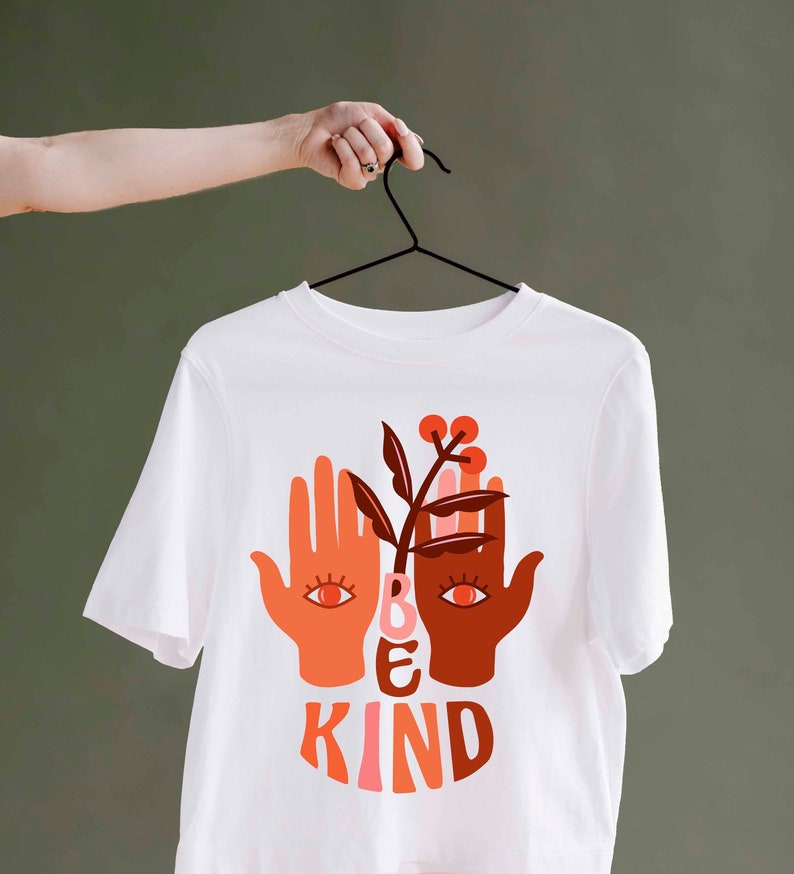 May include: White t-shirt with a graphic design featuring two hands with eyes, a plant, and the words "BE KIND" in orange and brown. The shirt is hanging on a black hanger.