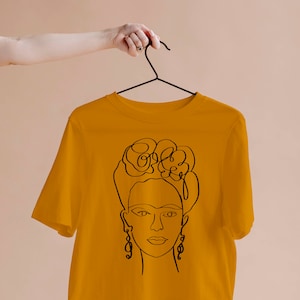 May include: Mustard yellow t-shirt featuring a black line drawing of a woman's face with floral headpiece and earrings. The shirt is hanging on a black hanger. The design is a minimalist, artistic representation.