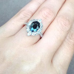 May include: A silver ring with a large, oval-cut, dark teal gemstone and a halo of smaller, clear stones. The ring's band is also set with small, clear stones. The ring is displayed against a neutral background.