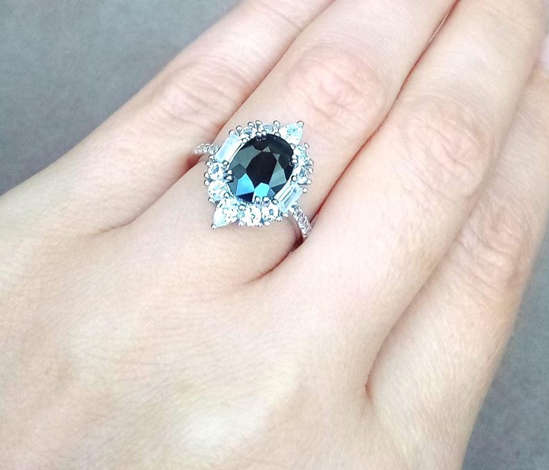 May include: A silver ring with a large, oval-cut, dark teal gemstone and a halo of smaller, clear stones. The ring's band is also set with small, clear stones. The ring is displayed against a neutral background.