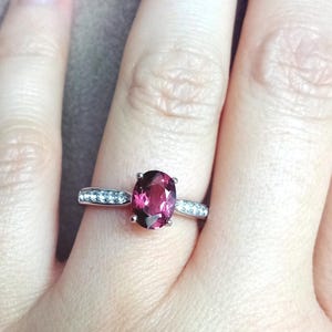 May include: A silver-toned ring featuring a central oval-cut deep pink gemstone held by four prongs. The band is accented with a row of small, clear, round-cut stones embedded along each side of the shank, creating a sparkling, refined appearance.