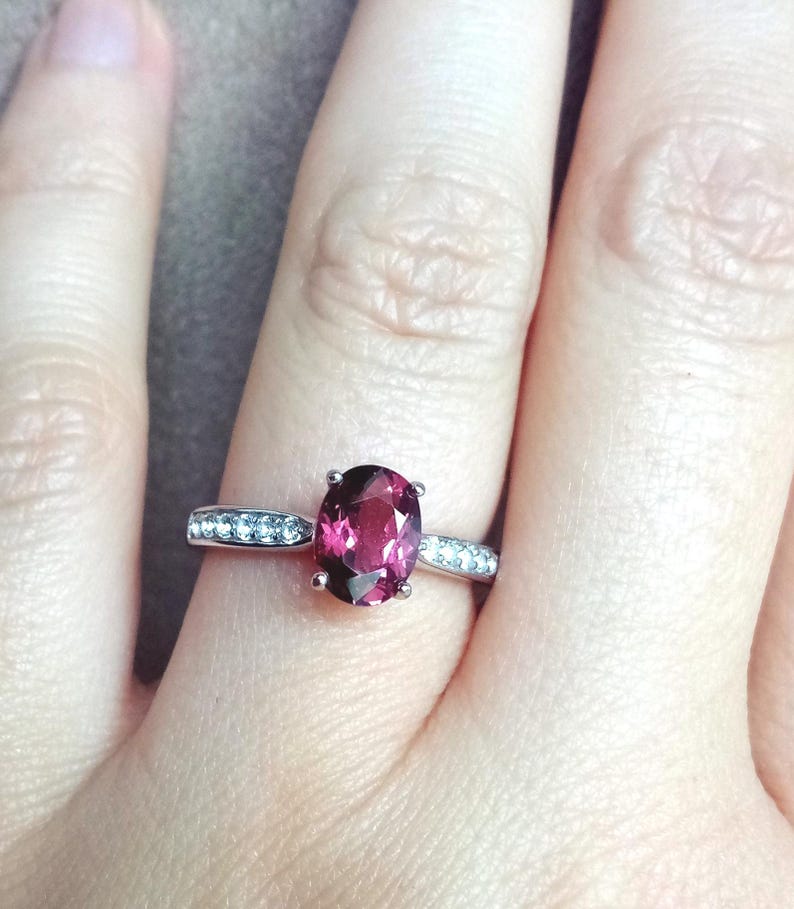 May include: A silver-toned ring featuring a central oval-cut deep pink gemstone held by four prongs. The band is accented with a row of small, clear, round-cut stones embedded along each side of the shank, creating a sparkling, refined appearance.