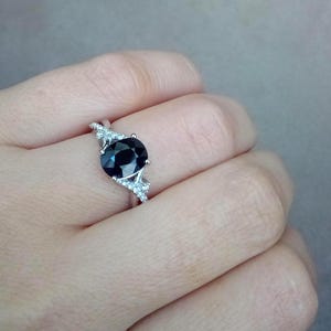 Puede incluir: Un anillo plateado con una gran piedra preciosa redonda de color azul oscuro como pieza central. El anillo est&aacute; adornado con piedras preciosas m&aacute;s peque&ntilde;as y claras a ambos lados de la piedra principal, creando un efecto brillante. El anillo se muestra en un dedo.