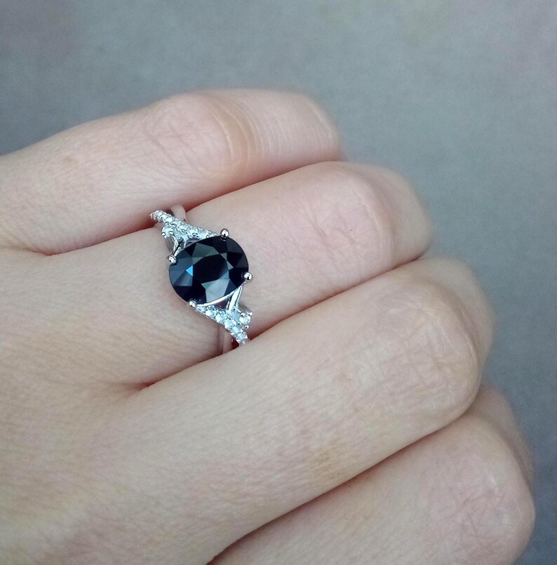 Puede incluir: Un anillo plateado con una gran piedra preciosa redonda de color azul oscuro como pieza central. El anillo est&aacute; adornado con piedras preciosas m&aacute;s peque&ntilde;as y claras a ambos lados de la piedra principal, creando un efecto brillante. El anillo se muestra en un dedo.