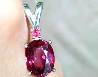 May include: A silver pendant featuring a large, oval-cut, deep pink gemstone held by four prongs. Above the main stone, a smaller, round, bright pink gemstone is set into the silver bail, which connects to a thin, light-colored string.