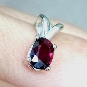 May include: A silver pendant with a teardrop-shaped, deep red gemstone. The gemstone is faceted, catching the light and displaying a range of dark red hues. The pendant has a simple, elegant design, suitable for everyday wear or special occasions.