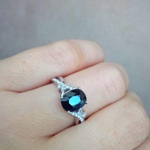Puede incluir: Un anillo de plata con una gran piedra preciosa ovalada de color azul oscuro. La banda est&aacute; adornada con piedras de acento m&aacute;s peque&ntilde;as y claras. El anillo se presenta sobre un fondo neutro, resaltando el dise&ntilde;o y el color de la joya.