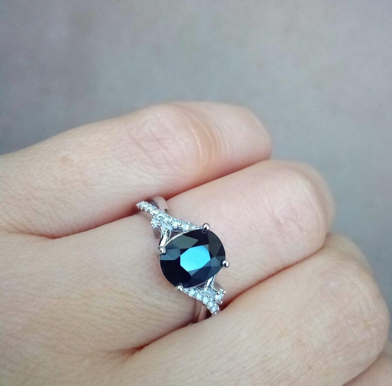 Puede incluir: Un anillo de plata con una gran piedra preciosa ovalada de color azul oscuro. La banda est&aacute; adornada con piedras de acento m&aacute;s peque&ntilde;as y claras. El anillo se presenta sobre un fondo neutro, resaltando el dise&ntilde;o y el color de la joya.