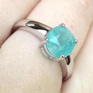 May include: A silver-toned metal ring featuring a round, faceted, bright turquoise-colored gemstone set in a four-prong setting. The band is smooth and polished, showcasing a reflective surface that catches the light around the central stone.