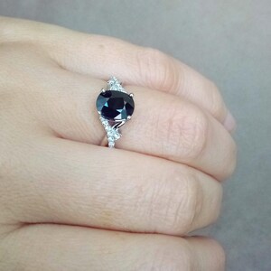 Puede incluir: Un anillo de plata con una gran piedra preciosa ovalada de color azul oscuro. El anillo est&aacute; adornado con piedras preciosas m&aacute;s peque&ntilde;as y claras en la banda, creando un efecto brillante. El anillo se muestra en el dedo de una persona.