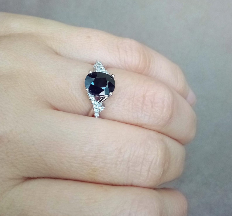 Puede incluir: Un anillo de plata con una gran piedra preciosa ovalada de color azul oscuro. El anillo est&aacute; adornado con piedras preciosas m&aacute;s peque&ntilde;as y claras en la banda, creando un efecto brillante. El anillo se muestra en el dedo de una persona.
