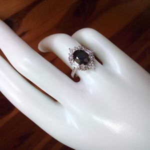 May include: A silver ring featuring a dark, oval-shaped gemstone, encircled by smaller, clear stones. The ring is presented on a white hand, with a wooden backdrop. The ring's design is detailed and sophisticated.