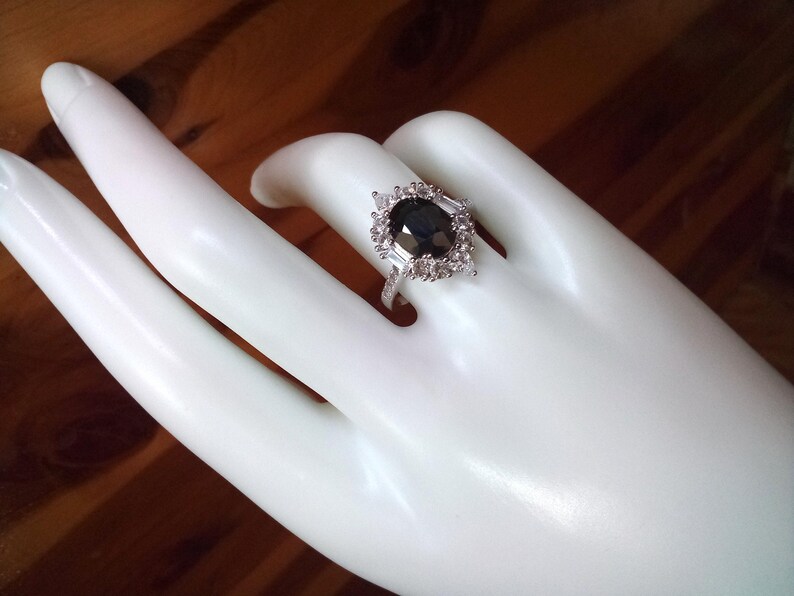 May include: A silver ring featuring a dark, oval-shaped gemstone, encircled by smaller, clear stones. The ring is presented on a white hand, with a wooden backdrop. The ring's design is detailed and sophisticated.