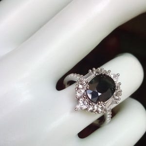 May include: A silver ring with a large, dark central gemstone, encircled by smaller, clear accent stones. The ring's design incorporates a halo of smaller stones and baguette-cut accents, producing a sparkling effect. The ring is shown on a white surface.
