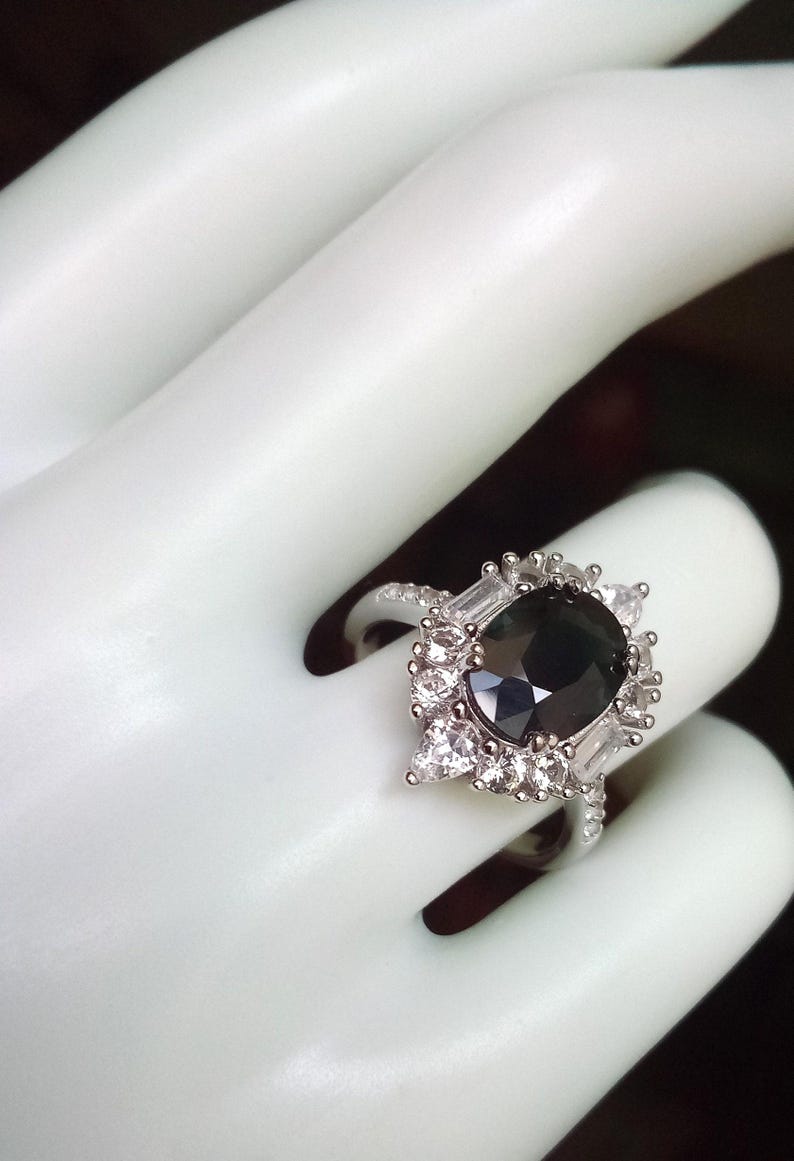 May include: A silver ring with a large, dark central gemstone, encircled by smaller, clear accent stones. The ring's design incorporates a halo of smaller stones and baguette-cut accents, producing a sparkling effect. The ring is shown on a white surface.
