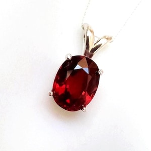 May include: A pendant necklace featuring a deep red, oval-cut gemstone secured in a four-prong silver-toned metal setting. The faceted stone displays a vibrant crimson hue, suspended from a delicate, thin silver-toned chain against a plain background.