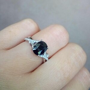 Puede incluir: Anillo plateado con una gran piedra negra de corte ovalado. La banda est&aacute; adornada con piedras m&aacute;s peque&ntilde;as y claras a ambos lados de la piedra principal, creando un efecto brillante. El anillo se presenta sobre un fondo neutro.