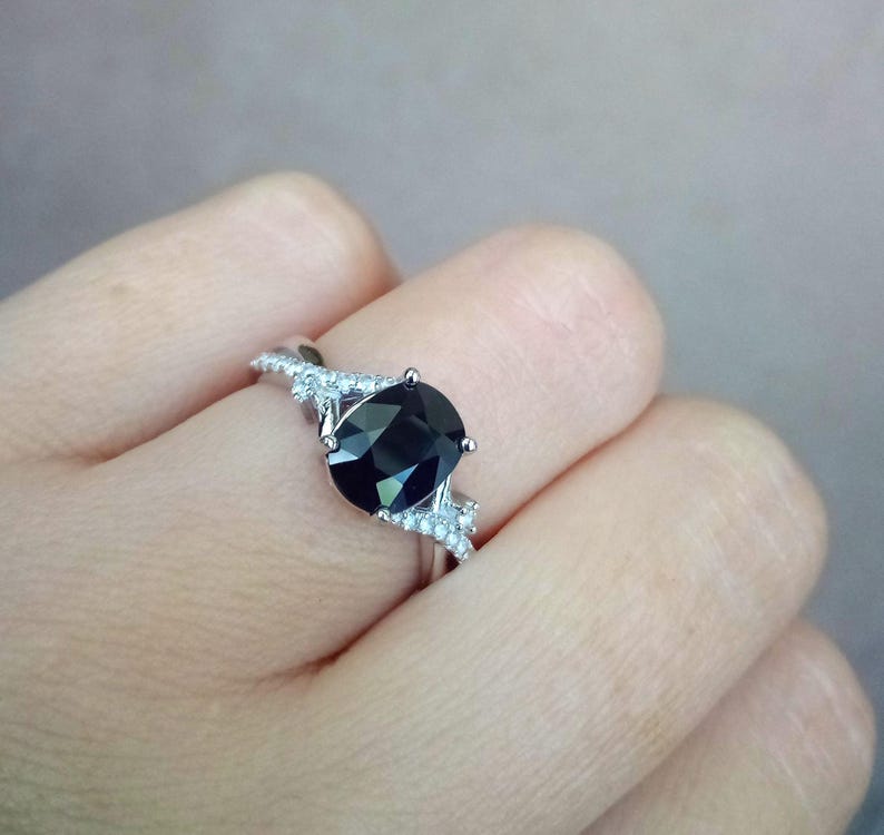 Puede incluir: Anillo plateado con una gran piedra negra de corte ovalado. La banda est&aacute; adornada con piedras m&aacute;s peque&ntilde;as y claras a ambos lados de la piedra principal, creando un efecto brillante. El anillo se presenta sobre un fondo neutro.
