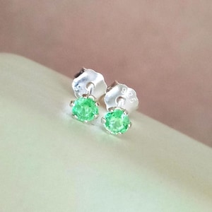 May include: A pair of stud earrings featuring round, vibrant green gemstones set in silver prongs. Each earring includes a silver post back clearly stamped with the number 925, indicating sterling silver construction for these small, circular jewelry pieces.