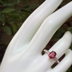 May include: A silver ring featuring a central oval-cut deep pink gemstone held by four prongs. The band is crafted from polished silver and is accented with a row of small, clear, round-cut stones embedded along each side of the central setting.