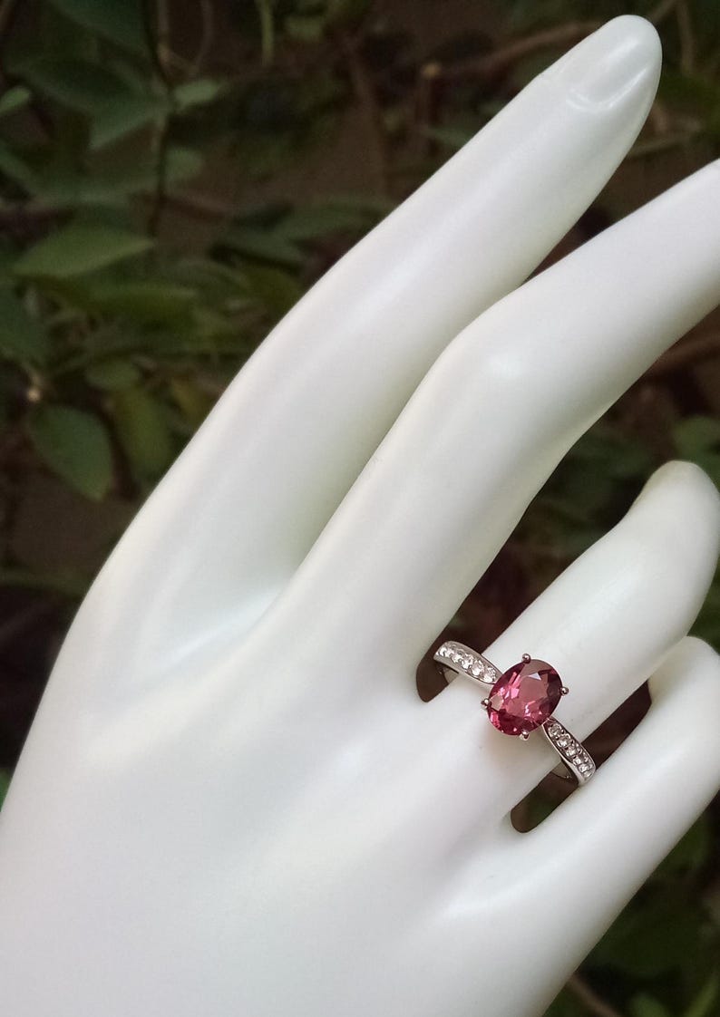 May include: A silver ring featuring a central oval-cut deep pink gemstone held by four prongs. The band is crafted from polished silver and is accented with a row of small, clear, round-cut stones embedded along each side of the central setting.
