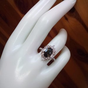 May include: A silver ring with a large, oval black gemstone, encircled by smaller, clear stones. The ring has a halo design with additional clear stones on the band, creating a sparkling effect. The ring is displayed on a white surface.