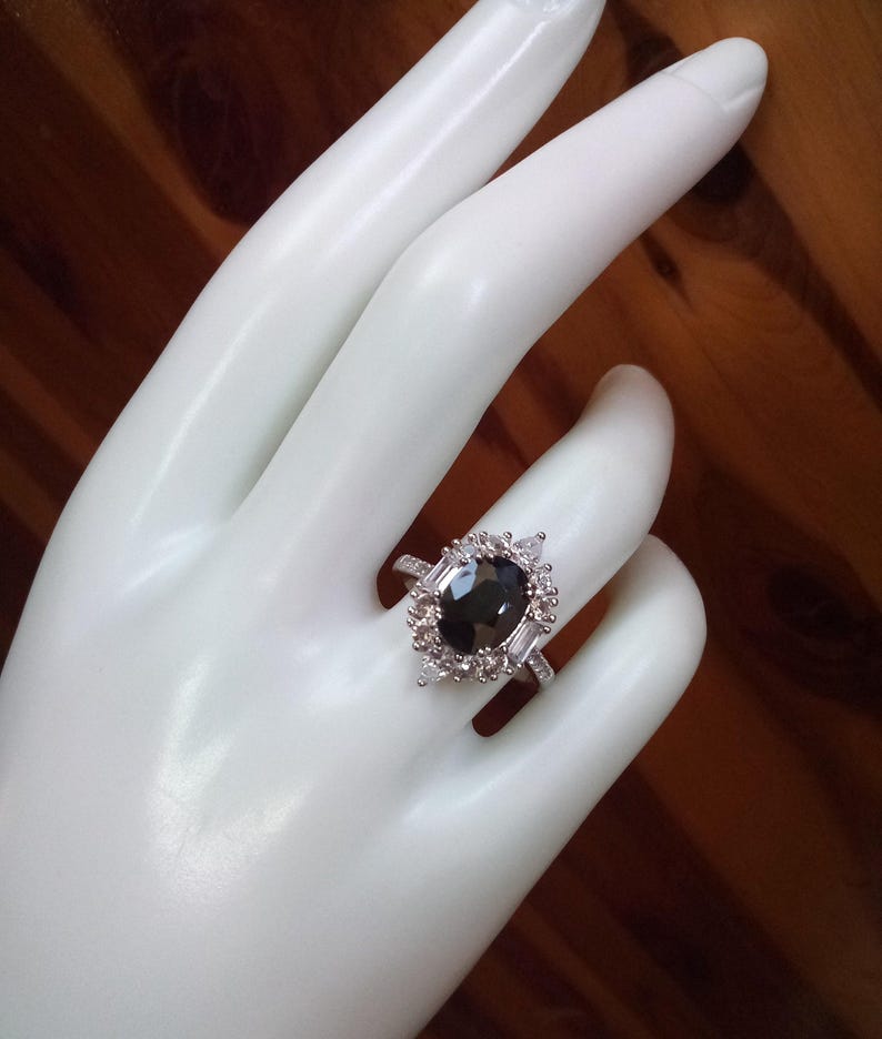 May include: A silver ring with a large, oval black gemstone, encircled by smaller, clear stones. The ring has a halo design with additional clear stones on the band, creating a sparkling effect. The ring is displayed on a white surface.
