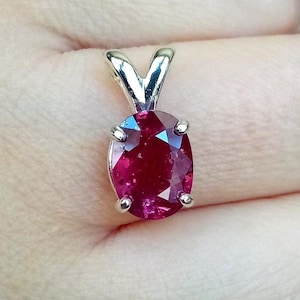 May include: An oval-cut deep red gemstone pendant set in a silver-toned metal prong setting. The faceted stone features a vibrant crimson hue with internal clarity characteristics, held securely by four metal prongs and a triangular bail.