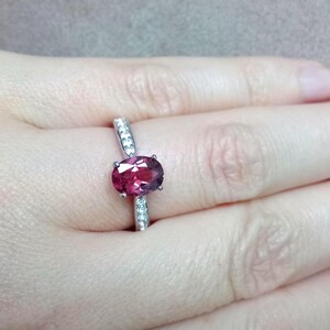 May include: A silver-toned ring featuring a central oval-cut deep pink gemstone held by four prongs. The band is accented with small, clear, round-cut stones embedded along the shank on both sides of the primary oval gemstone setting.