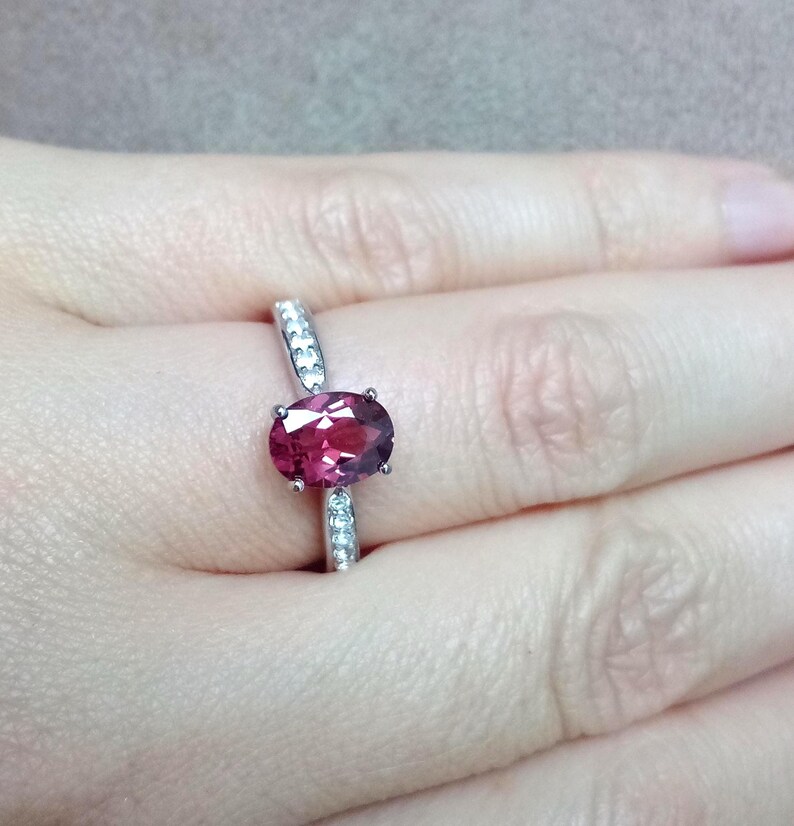 May include: A silver-toned ring featuring a central oval-cut deep pink gemstone held by four prongs. The band is accented with small, clear, round-cut stones embedded along the shank on both sides of the primary oval gemstone setting.