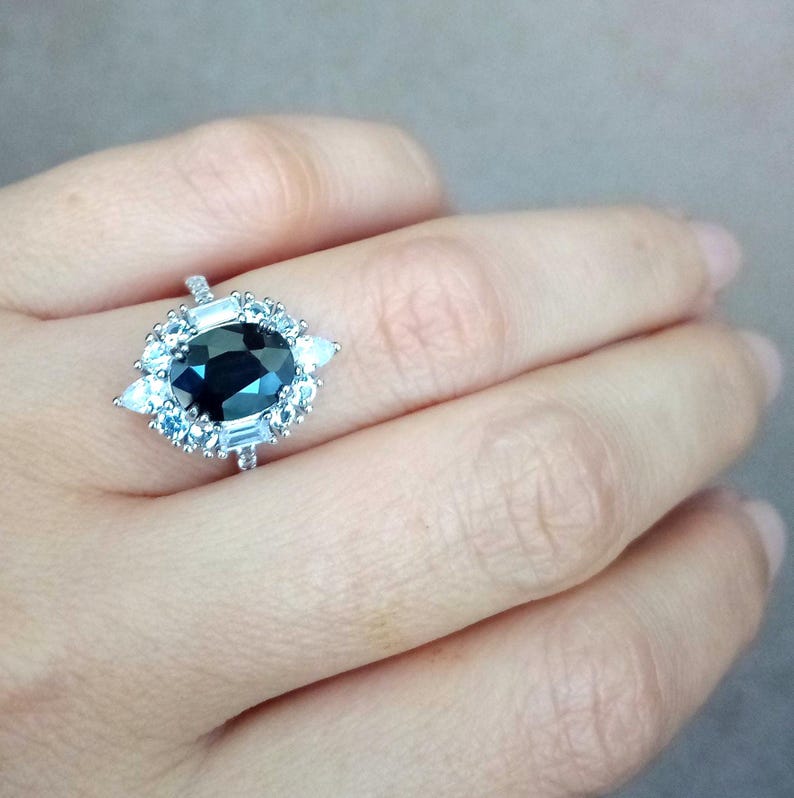 May include: A silver ring with a large, dark blue oval gemstone, encircled by smaller light blue and white stones. The ring has a halo design with baguette-cut stones. The ring is on a person's finger.